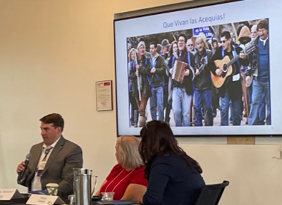 panelists seated at table in front of photo of acequias advocates